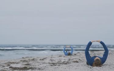 2 pessoas deitadas na areia da praia, vestidas, de barriga para cima com os braços em 1ª posição de dança